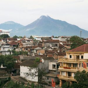 Town with tiled rooftops and a yellow building beneath a large volcano, with mountains and cloudy sky beyond